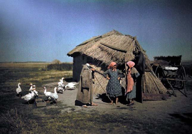 Women stand in front of a primitive straw covered hut