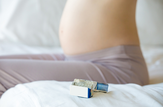 Closeup of torso of young smoker pregnant woman sitting on the bed. Focus on pack of cigarettes